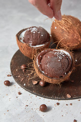 Female hand coconut sprinkles on chocolate ice cream in coconut halves on a gray stone background.