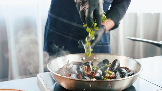 Chef sprinkles a parsley on mussels in frying pan, close-up in slow motion.