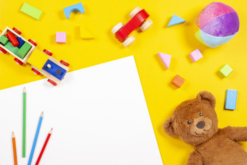 Baby kids toys background. Blank white card, colored pencils, wooden and soft toys on yellow background. Top view