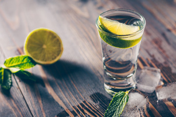 Cold vodka in a shot glass on a wooden table with a slice of lime, mint and ice slices
