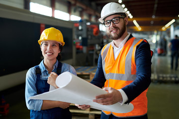 Portrait of modern bearded engineer wearing hardhat holding blueprints smiling cheerfully while discussing production with female worker at factory, copy space