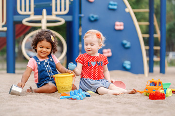 Summer mood. Two cute Caucasian and hispanic latin babies children sitting in sandbox playing with plastic colorful toys. Little girls friends having fun together on playground.