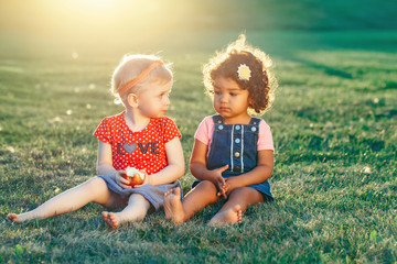 Group portrait of white Caucasian and latin hispanic girls children sitting together sharing apple. Two babies eating fruits outside in park on summer day. Best friends forever. Healthy childhood.