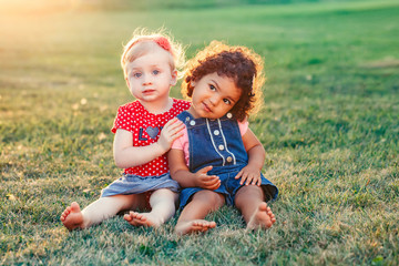 Fototapeta premium Group portrait of two cute adorable girls toddlers children sitting together. White Caucasian and latin hispanic babies hugging outside in park. Friendship and best friends forever concept.