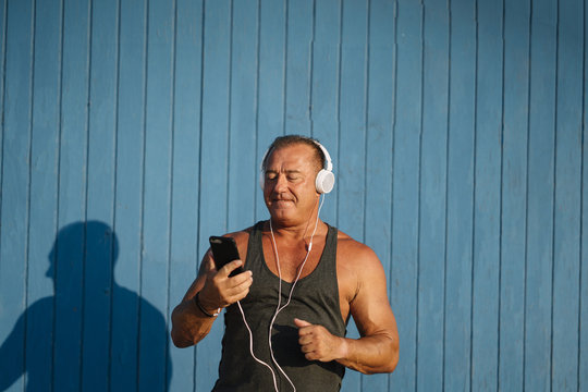 Strong older man poses with headphones on blue cabin background.