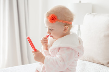 Cute asorable funny Caucasian blonde baby girl 2 years old in bathrobe doing applying makeup, looking into mirror and sitting on her bed in bedroom at home. Happy childhood concept.