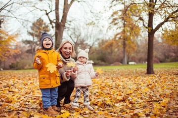Fototapeta premium A young beautiful mother and two small children are walking around the autumn park. Mom and two small children play. Warm winter. Bright autumn.
