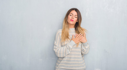 Fototapeta premium Young adult woman over grunge grey wall wearing winter sweater smiling with hands on chest with closed eyes and grateful gesture on face. Health concept.