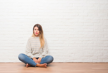 Young adult woman sitting on the floor over white brick wall at home skeptic and nervous, frowning upset because of problem. Negative person.