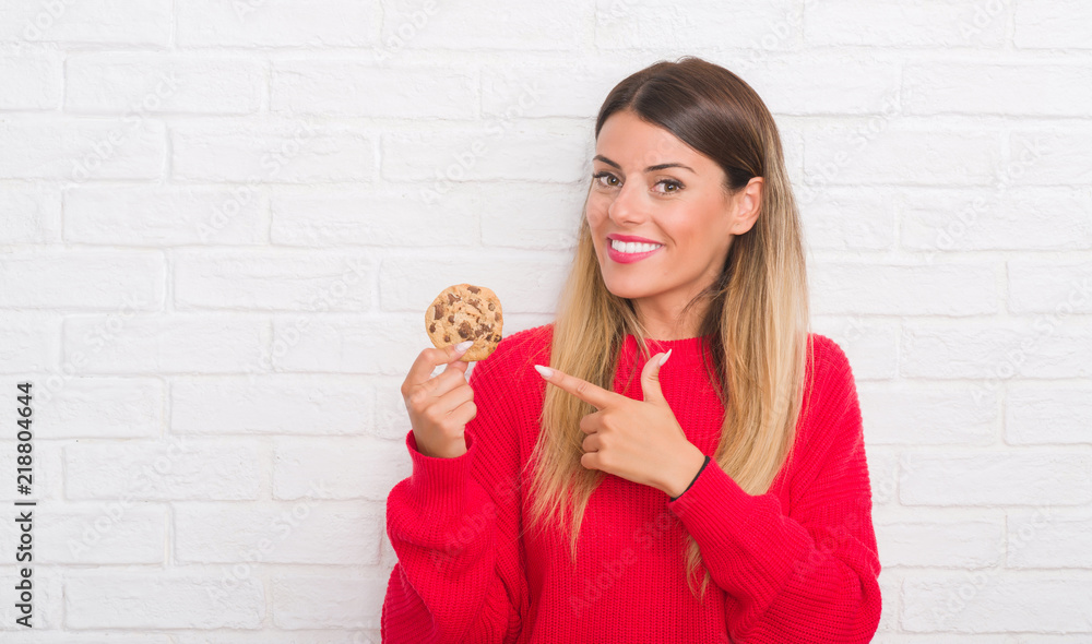 Poster young adult woman over white brick wall eating chocolate chip cooky very happy pointing with hand an