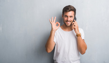 Fototapeta premium Handsome young man over grey grunge wall speaking on the phone doing ok sign with fingers, excellent symbol