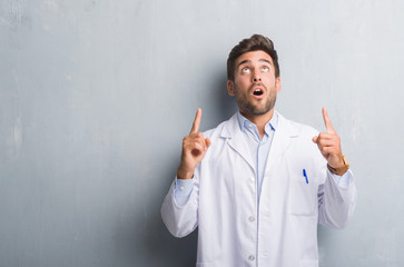 Handsome young professional man over grey grunge wall wearing white coat amazed and surprised looking up and pointing with fingers and raised arms.