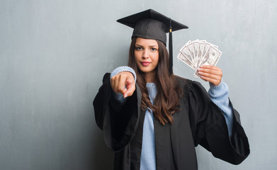 Young brunette woman over grunge grey wall wearing graduate uniform holding dollars pointing with finger to the camera and to you, hand sign, positive and confident gesture from the front