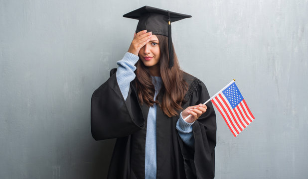 Young brunette woman over grunge grey wall wearing graduate uniform holding flag of America stressed with hand on head, shocked with shame and surprise face, angry and frustrated. Fear and upset