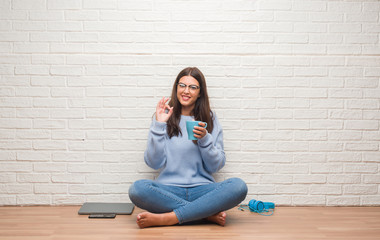 Young brunette woman sitting on the floor over white brick wall drinking coffee doing ok sign with fingers, excellent symbol