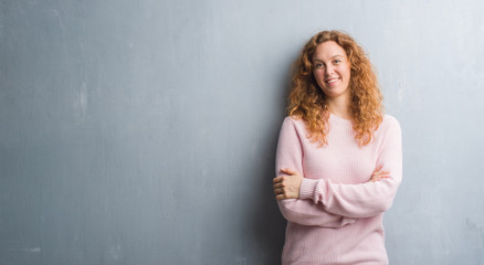 Young redhead woman over grey grunge wall wearing pink sweater happy face smiling with crossed arms...