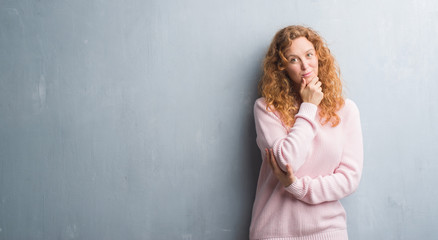 Fototapeta premium Young redhead woman over grey grunge wall wearing pink sweater looking confident at the camera with smile with crossed arms and hand raised on chin. Thinking positive.