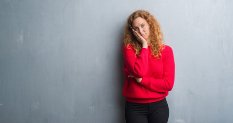 Young redhead woman over grey grunge wall wearing red sweater thinking looking tired and bored with depression problems with crossed arms.