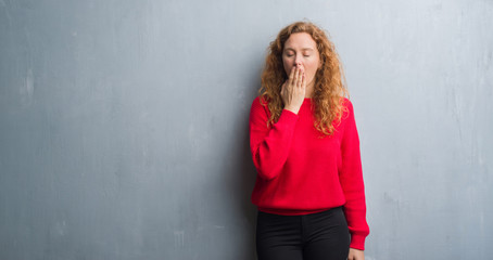 Young redhead woman over grey grunge wall wearing red sweater bored yawning tired covering mouth with hand. Restless and sleepiness.