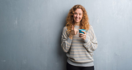 Young redhead woman over grey grunge wall drinking a cup of coffee happy with big smile doing ok sign, thumb up with fingers, excellent sign