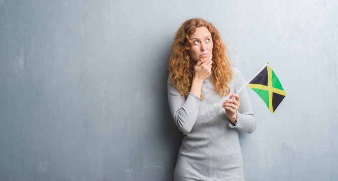 Young redhead woman over grey grunge wall holding flag of Jamaica serious face thinking about question, very confused idea