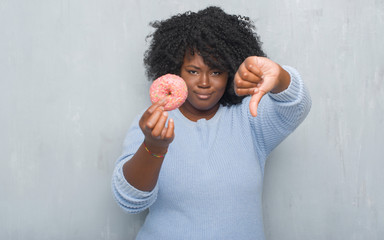 Young african american woman over grey grunge wall eating pink donut with angry face, negative sign...