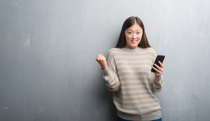 Young Chinese woman over grey wall looking at smartphone screaming proud and celebrating victory and success very excited, cheering emotion