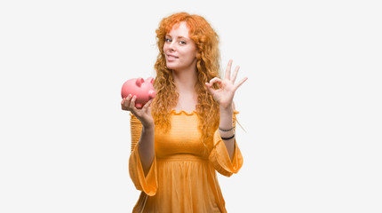Young redhead woman holding piggy bank doing ok sign with fingers, excellent symbol