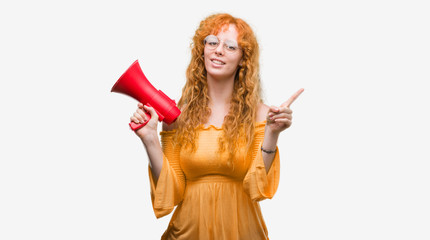 Young redhead woman holding megaphone very happy pointing with hand and finger to the side