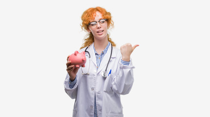 Young redhead doctor woman holding piggy bank pointing and showing with thumb up to the side with happy face smiling