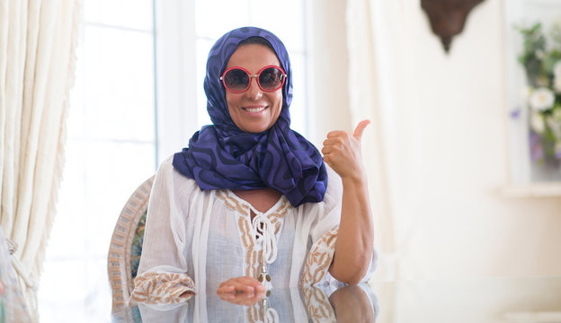 Middle age brunette arabian woman wearing colorful hijab at luxury house pointing and showing with thumb up to the side with happy face smiling