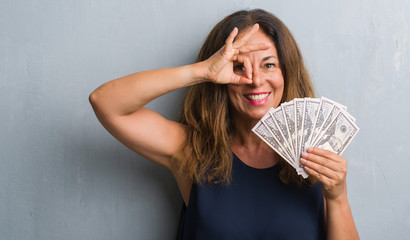 Middle age hispanic woman standing over grey grunge wall holding dollars with happy face smiling doing ok sign with hand on eye looking through fingers