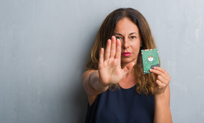 Middle age hispanic woman standing over grey grunge wall holding hard drive with open hand doing stop sign with serious and confident expression, defense gesture