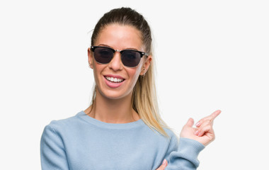 Beautiful young woman wearing sunglasses and ponytail very happy pointing with hand and finger to the side