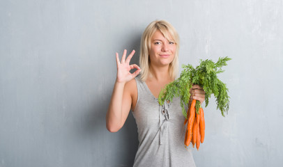 Caucasian adult woman over grey grunge wall holding fresh carrots doing ok sign with fingers,...