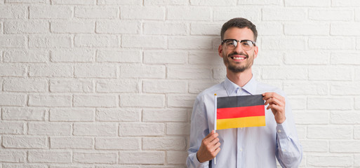 Young adult man over brick wall holding flag of Germany with a happy face standing and smiling with a confident smile showing teeth