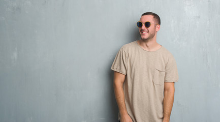 Young caucasian man over grey grunge wall wearing sunglasses looking away to side with smile on face, natural expression. Laughing confident.