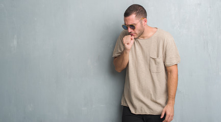 Young caucasian man over grey grunge wall wearing sunglasses feeling unwell and coughing as symptom for cold or bronchitis. Healthcare concept.