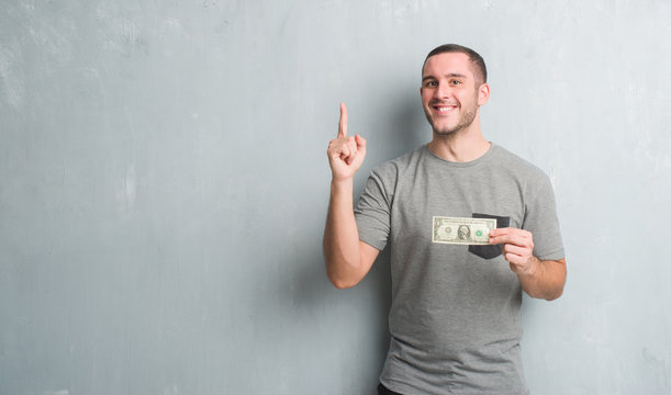 Young caucasian man over grey grunge wall showing one dollar surprised with an idea or question pointing finger with happy face, number one