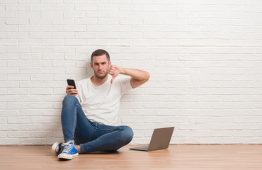 Young caucasian man sitting over white brick wall using computer laptop and smartphone with angry...