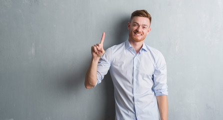 Young redhead business man over grey grunge wall showing and pointing up with finger number one...