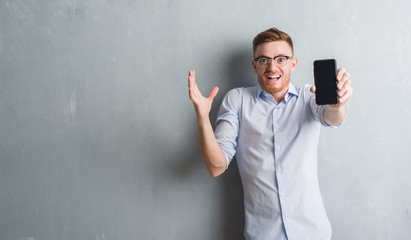 Young redhead man over grey grunge wall showing blank screen of smartphone very happy and excited, winner expression celebrating victory screaming with big smile and raised hands