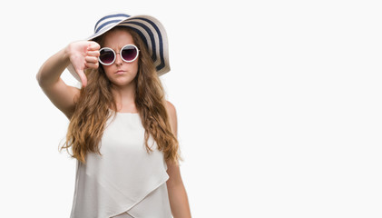 Young blonde woman wearing sunglasses and summer hat with angry face, negative sign showing dislike with thumbs down, rejection concept