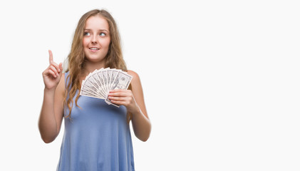 Obraz premium Young blonde woman holding dollars surprised with an idea or question pointing finger with happy face, number one