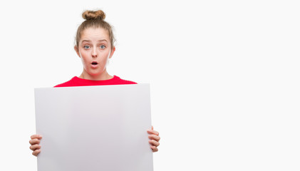 Young blonde woman holding advertising banner scared in shock with a surprise face, afraid and...