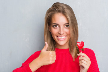 Beautiful young woman over grunge grey wall eating red heart lollipop candy happy with big smile doing ok sign, thumb up with fingers, excellent sign