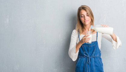 Beautiful young woman over grunge grey wall driking a glass of milk with a confident expression on...