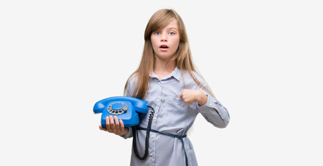Young blonde toddler holding vintage telephone with surprise face pointing finger to himself