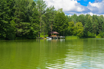 Obraz premium A beautiful image of a landscape from center of a river surrounded by trees and reeds on the shore against a blue sky in the clouds. Wooden gazebo on the beach. Reflection, water, tourist destination