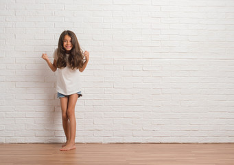 Young hispanic kid stading over white brick wall celebrating surprised and amazed for success with...